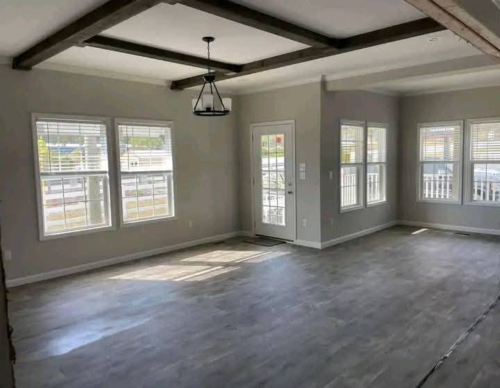Bright, unfurnished living room with large windows allowing natural light. Features wooden beams on the ceiling, a hanging light fixture, and light wood flooring.