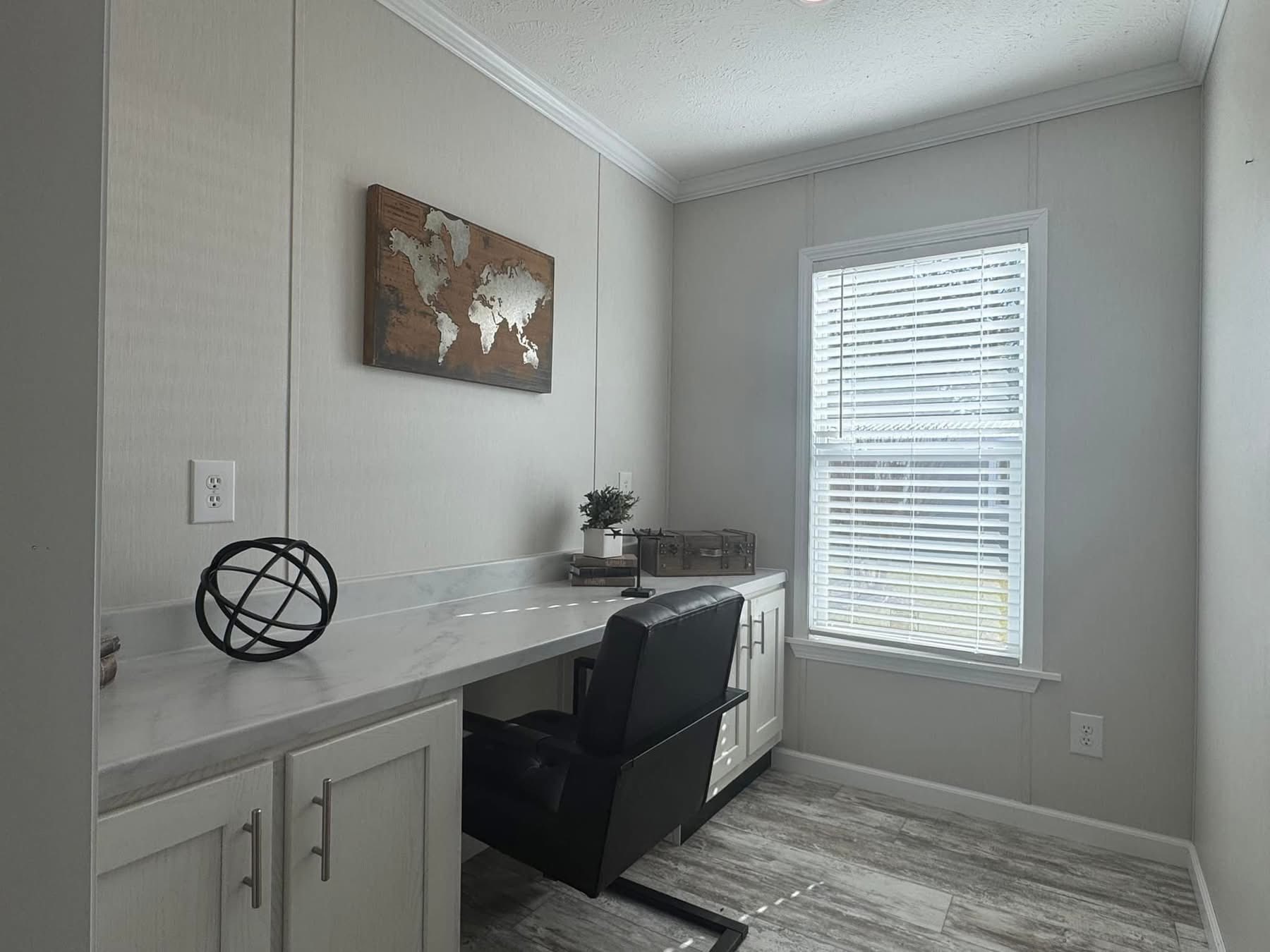 A small, tidy office with a sleek desk and black chair. A world map decorates the wall, and a window with blinds allows soft light. Neutral tones create a calm atmosphere.