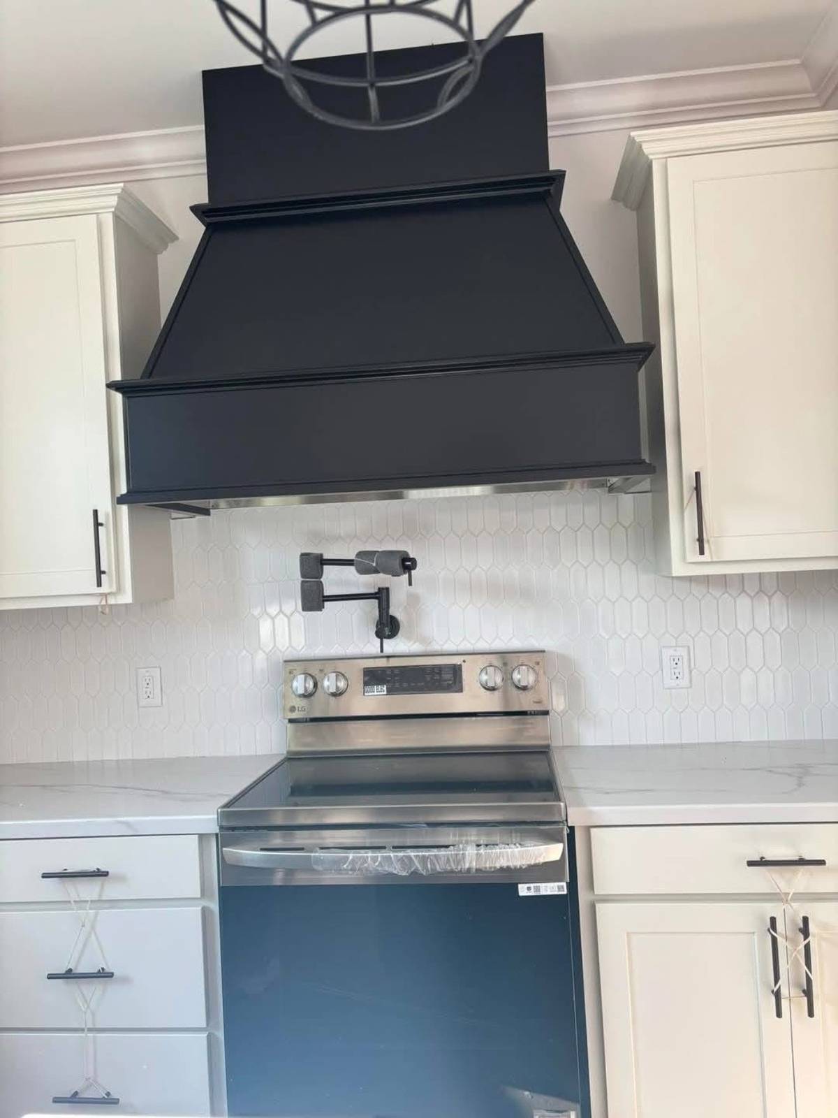 Modern kitchen with a stainless steel stove, black range hood, white hexagonal tile backsplash, and white cabinets. The space is sleek and clean.