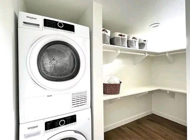 Laundry room with a stacked white washer and dryer on the left. The right features open shelves with gray baskets and a woven hamper, creating a tidy, modern look.