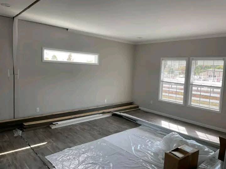 A room under renovation with light gray walls, new flooring partially installed, and stacked wood planks. Sunlight streams through two windows. Calm ambiance.