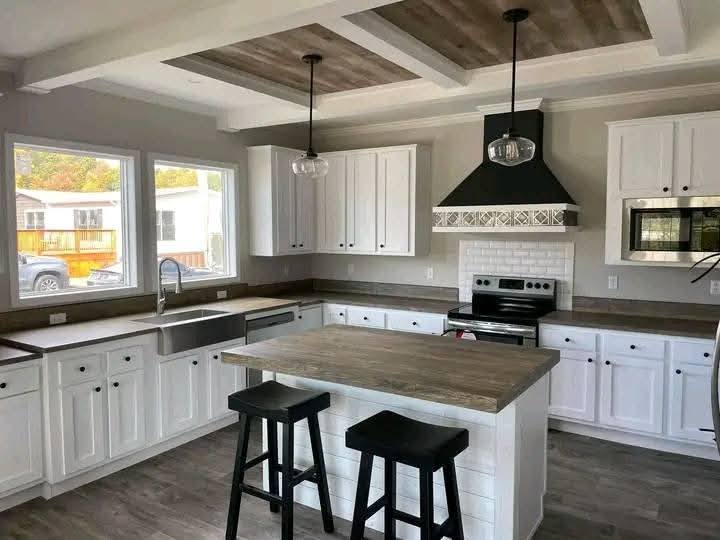 Bright, modern kitchen with white cabinets, stainless steel appliances, and wood accents. Features an island with two black stools, pendant lighting, and large windows.