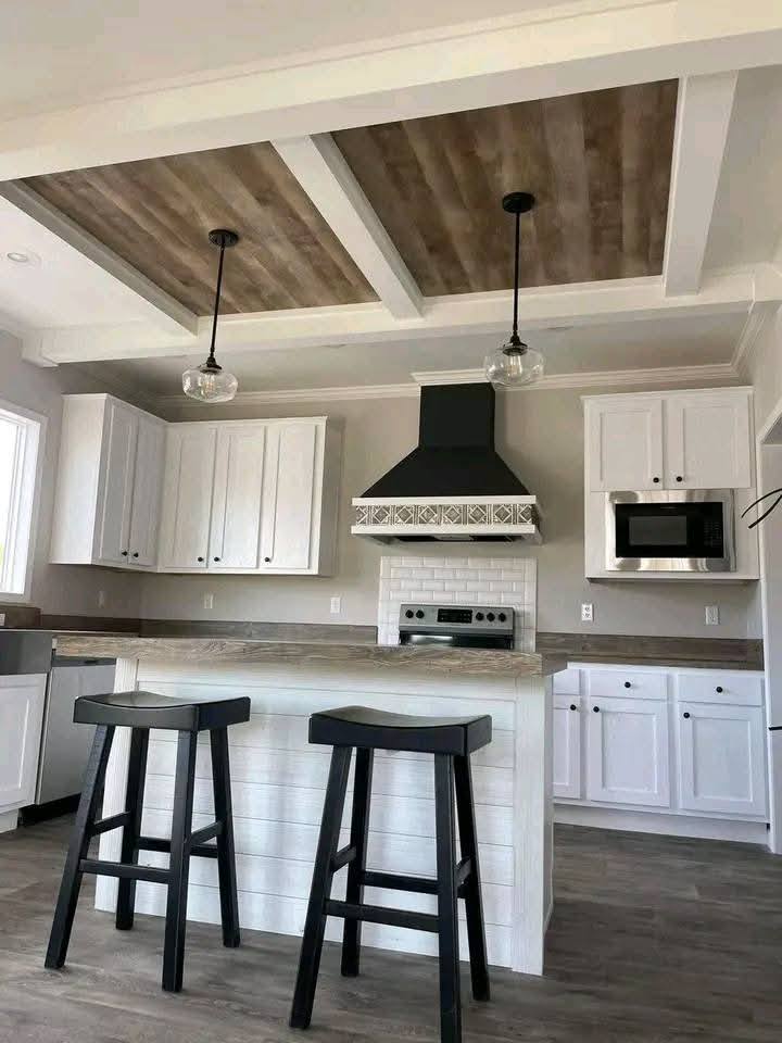 Modern kitchen with white cabinets, wood-accented ceiling, and island. Two black stools face the island; pendant lights hang above, creating a cozy atmosphere.