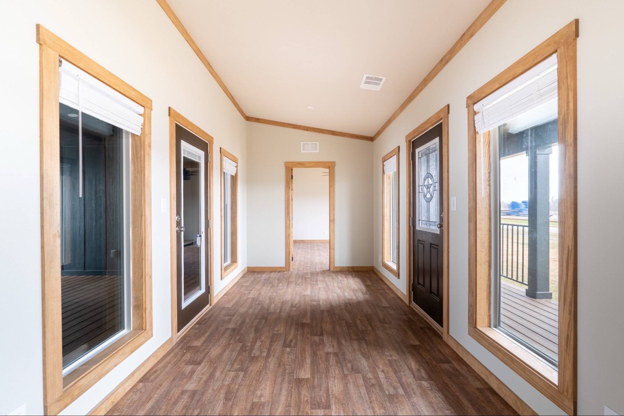 A bright hallway with beige walls and wood trim features large windows and glass doors on both sides. The wooden floor enhances the warm, welcoming feel.