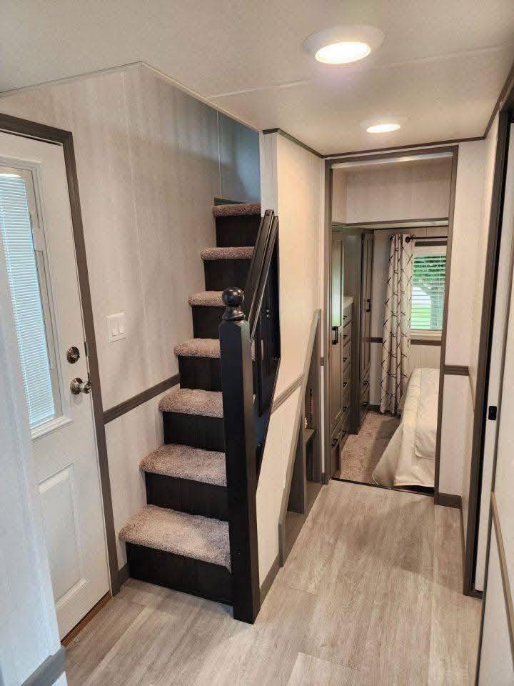 Narrow hallway with light wood flooring, carpeted staircase on the left, leading to a small bedroom with white bedding and a window with patterned curtains.