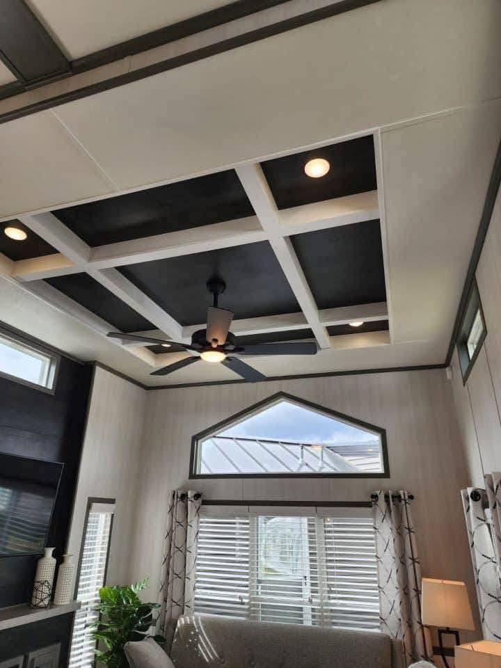 Modern living room with a coffered ceiling and recessed lighting. A ceiling fan hangs centrally. Tall windows and curtains accentuate natural light.