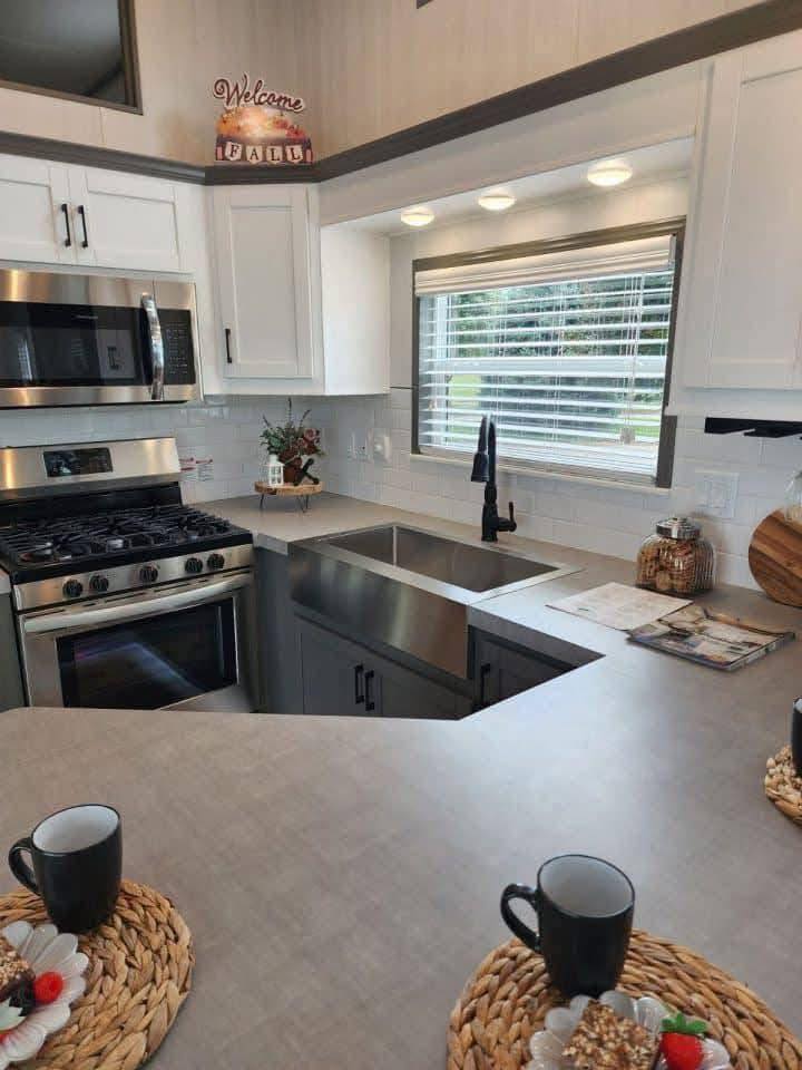 Modern kitchen with white cabinets, stainless steel appliances, and a dual sink under a bright window. Counter holds mugs on wicker placemats. Cozy fall decor.