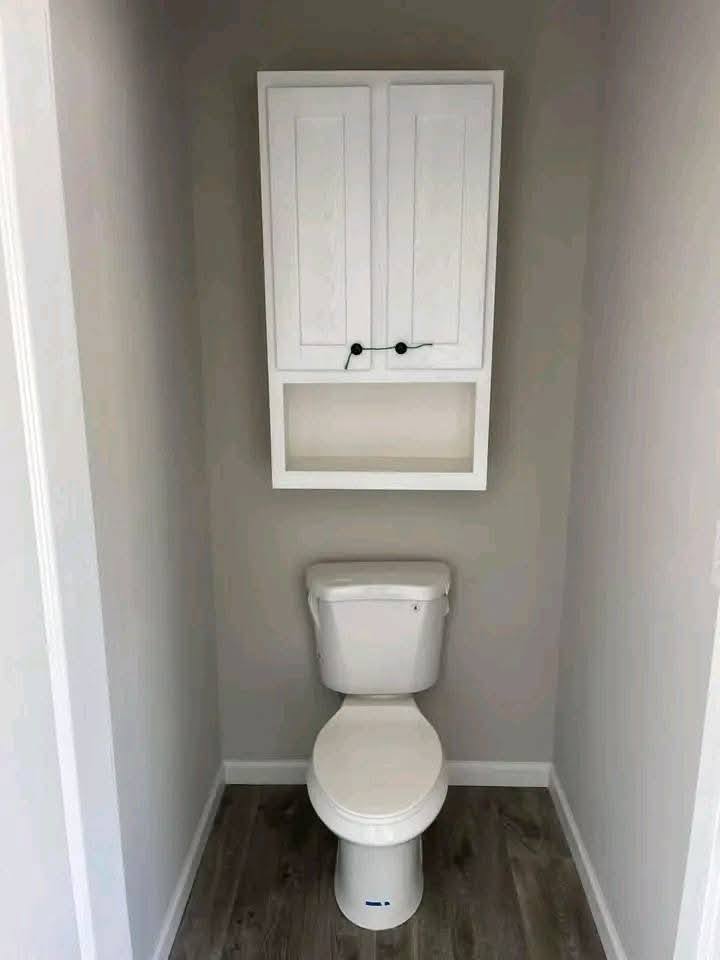 Small bathroom with a white toilet centered against a gray wall. Above it, a white cabinet with dark handles is mounted. The floor is gray wood.