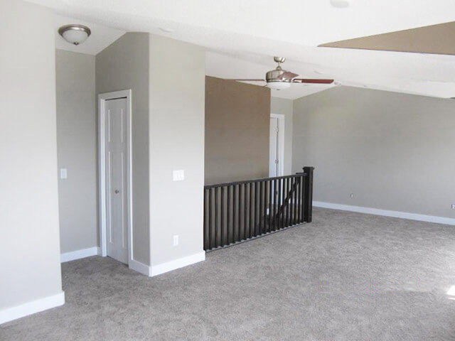 Bright, spacious room with gray walls and carpet, featuring a white door, dark railing, ceiling fan, and recessed lighting, conveying a modern, clean look.