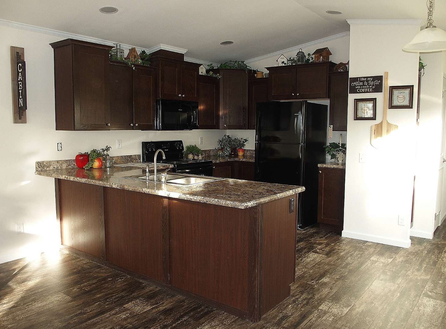 Warm kitchen with dark wood cabinets, granite countertops, and stainless steel sink. Black appliances add contrast. Decor includes greenery and rustic signs. Inviting feel.