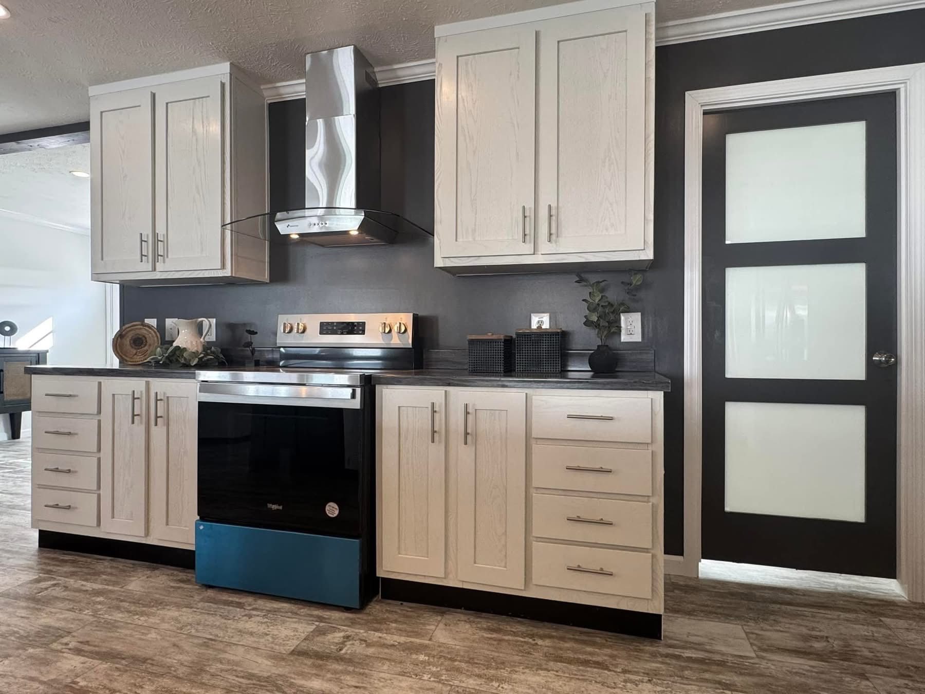 Modern kitchen with light wood cabinets, stainless steel appliances, and a glass range hood against a dark wall. A frosted glass door is on the right.