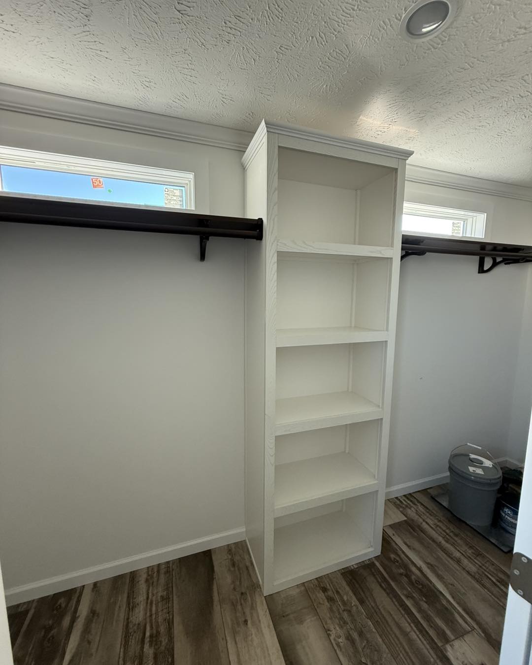 Minimalist wooden closet with light walls and ceiling, showcasing brown shelves and hanging rods. Sunlight filters through small windows, creating a cozy atmosphere.