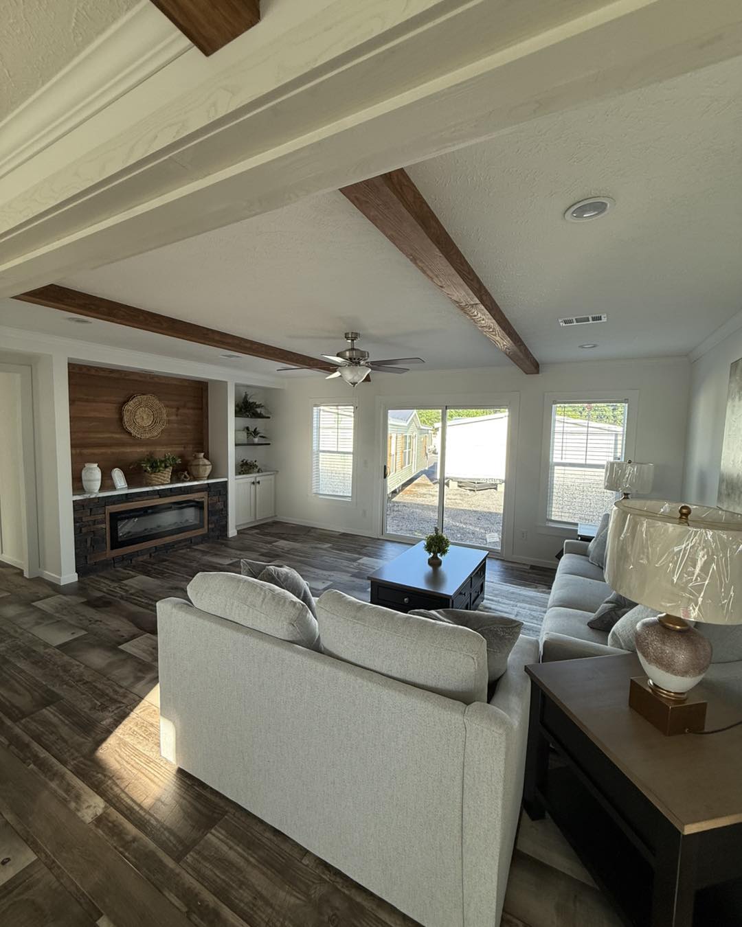 A cozy living room with light gray sofas, a wooden coffee table, and ceiling beams. A brick fireplace and large windows add warmth and natural light.