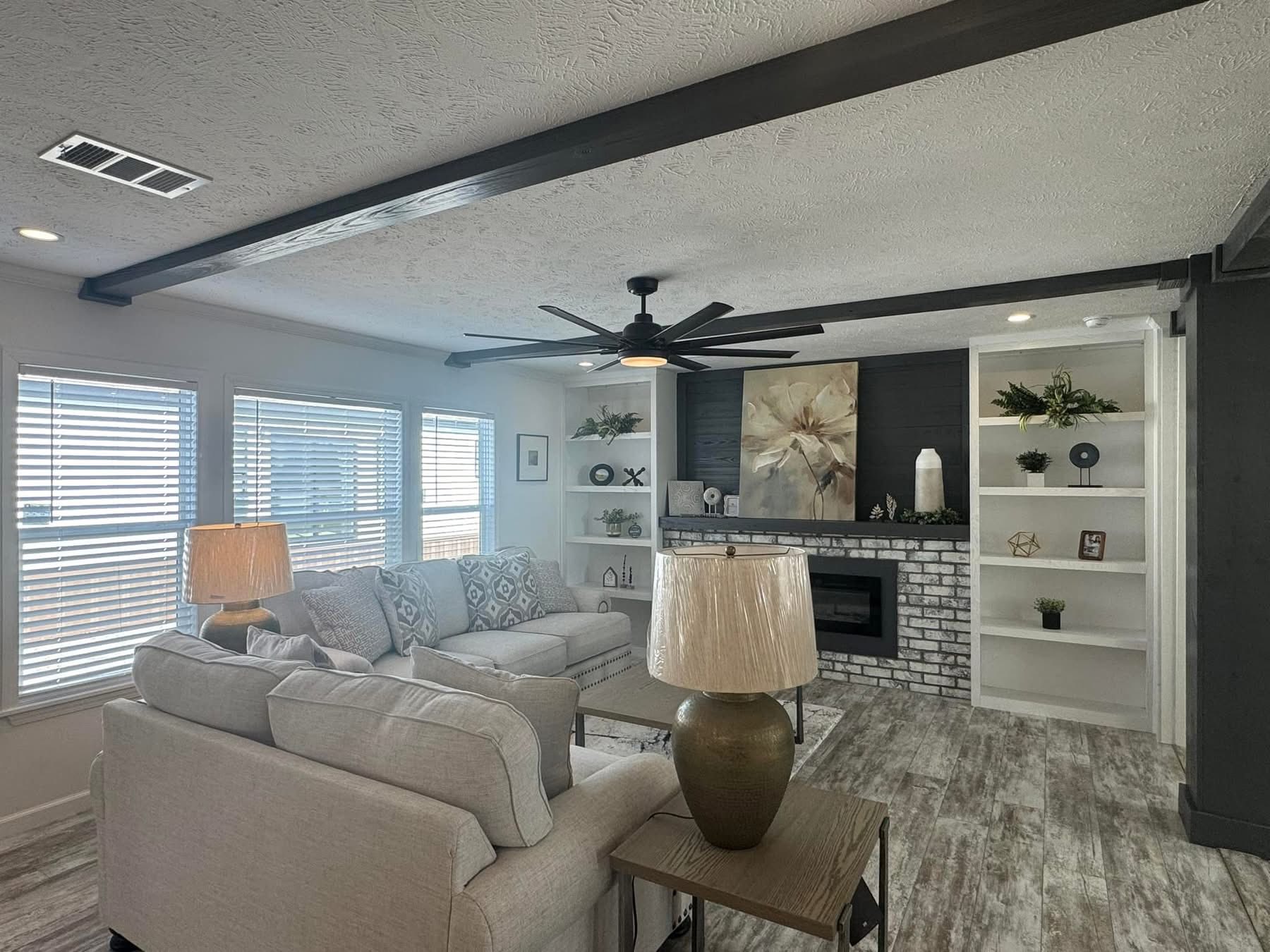 Elegant living room with gray sofas, patterned cushions, and a wooden floor. Features a white brick fireplace, built-in shelves, and a black ceiling fan. Cozy and modern ambiance.
