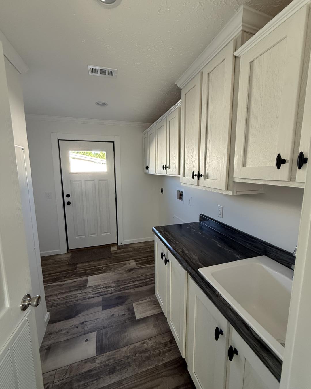 Narrow, well-lit utility room with white cabinets, dark countertops, and a sink. Wooden floor and door with window panels. Clean and organized.