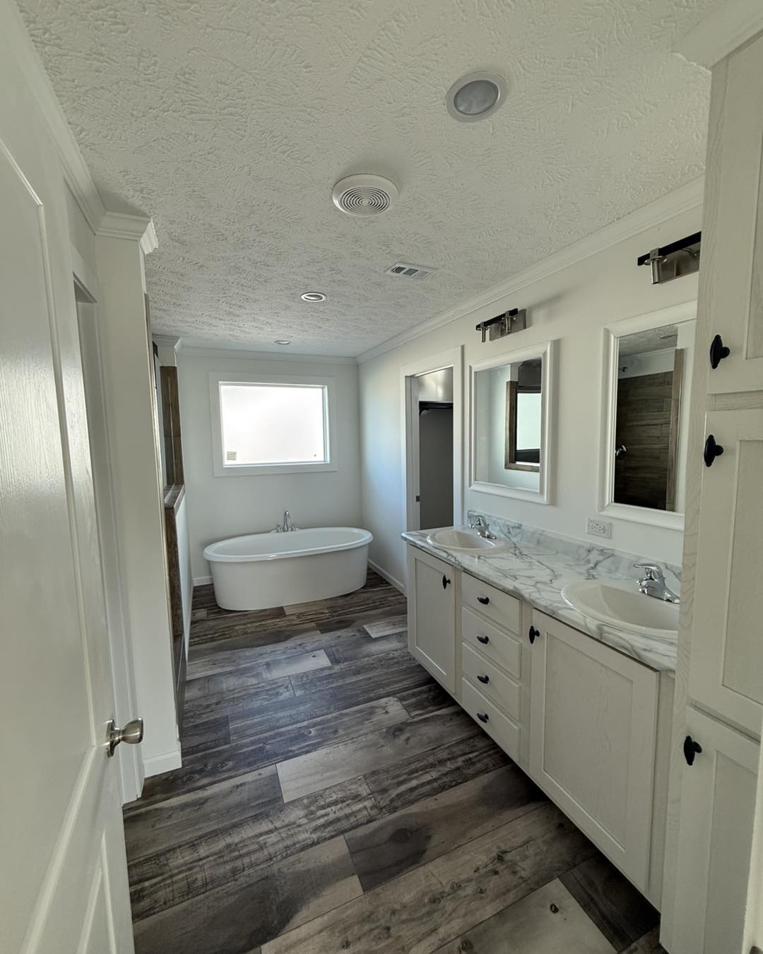 Modern bathroom with a freestanding bathtub under a frosted window. Dual marble sinks on a wooden vanity. Neutral tones and wood-style flooring.