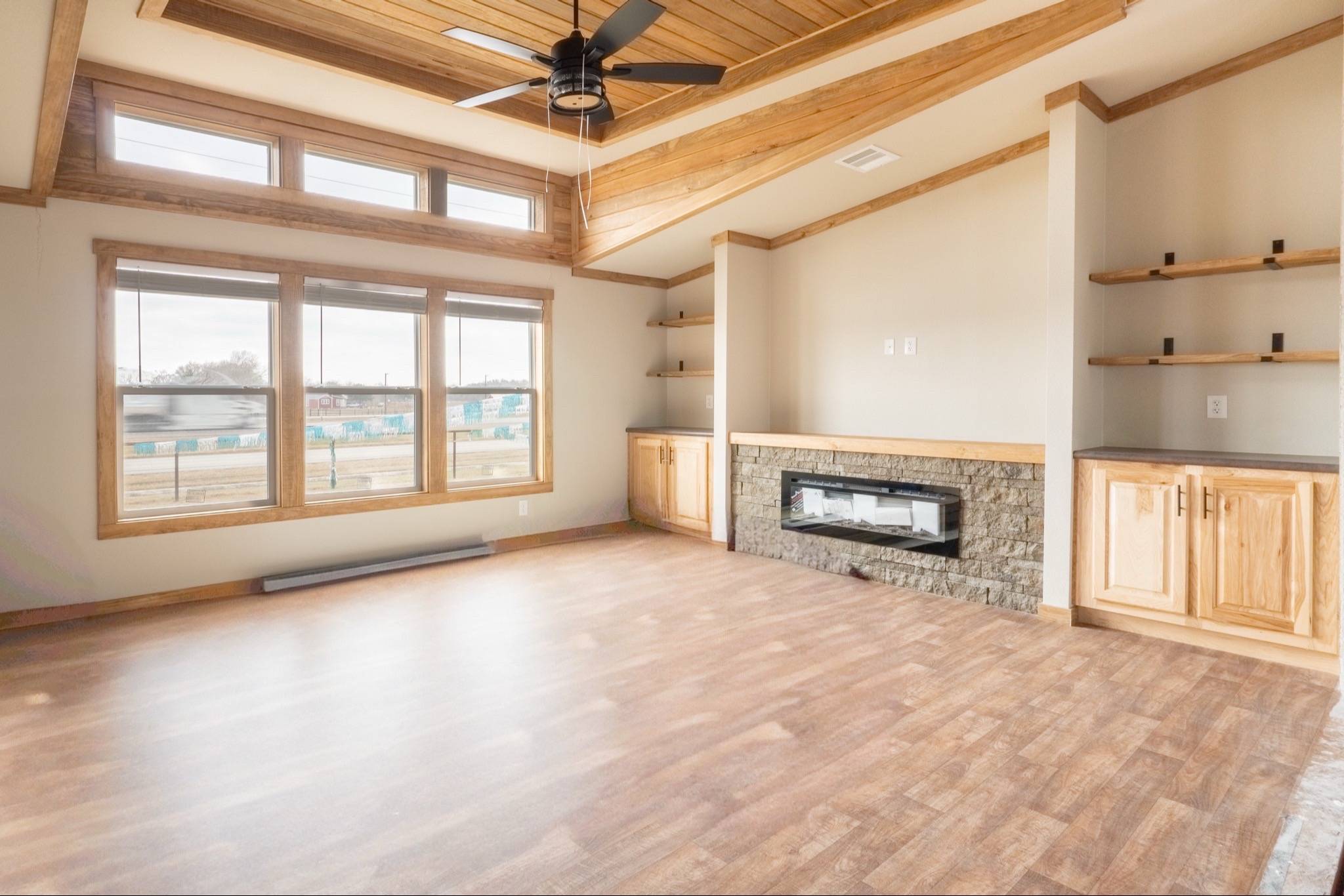 Spacious living room with hardwood floors, large windows, wood panel ceiling, modern fireplace, wooden shelves, and cabinets. Bright and airy feel.