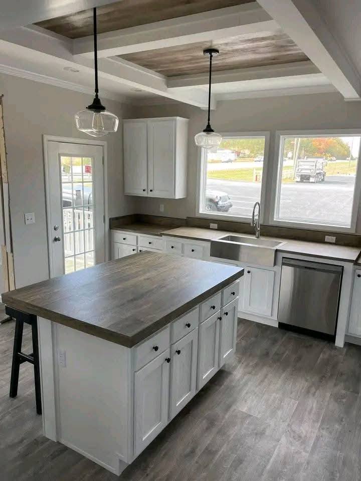 Bright kitchen with white cabinets, a large island, and wood countertops. Two pendant lights hang above. Windows offer a view of a sunny day.