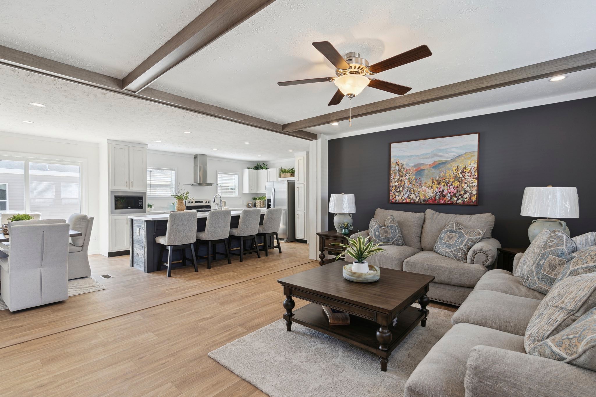 Spacious living room with beige sofas, wooden coffee table, and plant, adjacent to a dining area. Warm, modern tones with wood flooring and ceiling fan.