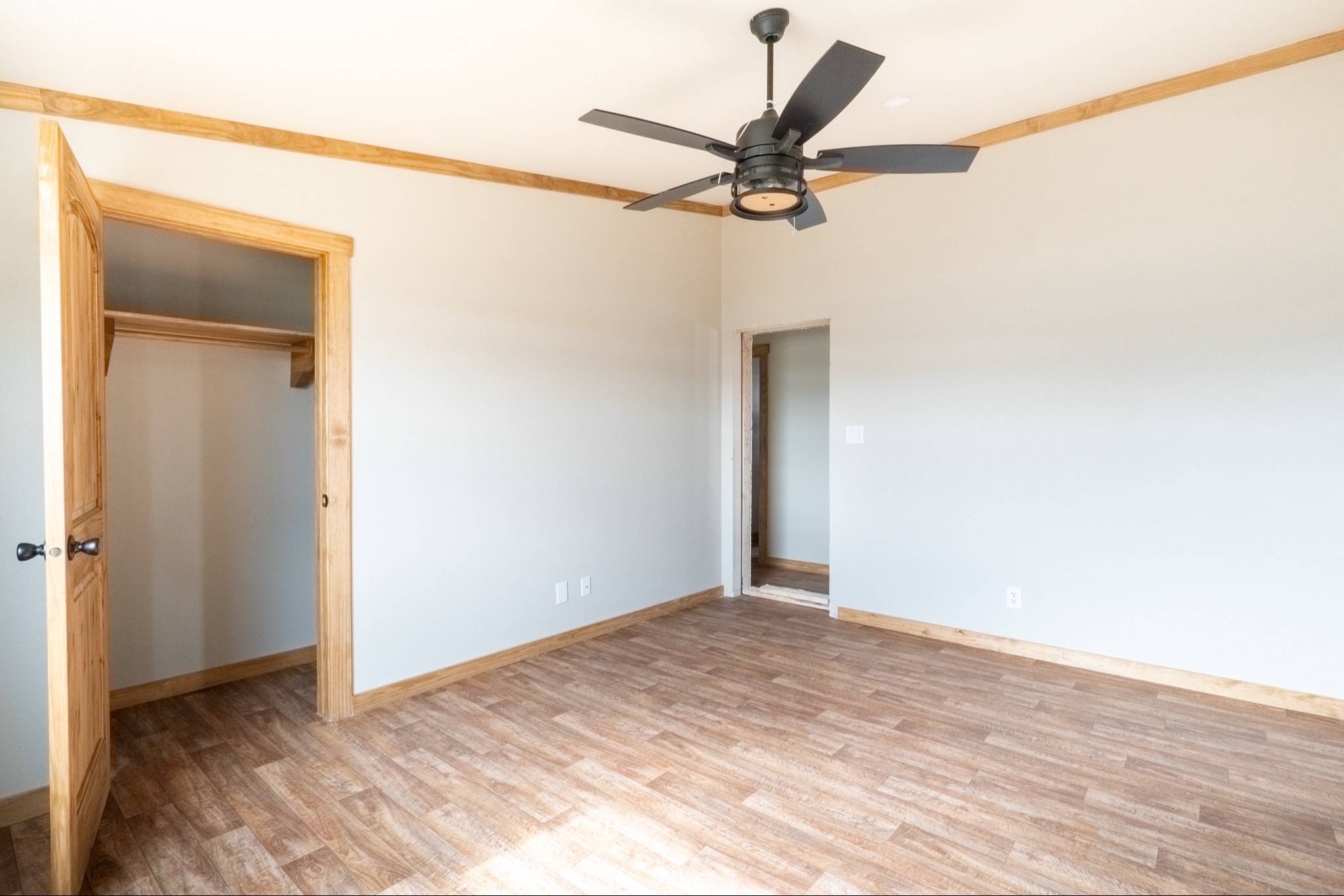 Empty room with wood flooring, beige walls, and ceiling fan. Open closet door on the left, hallway entrance on the right, creating a spacious feel.