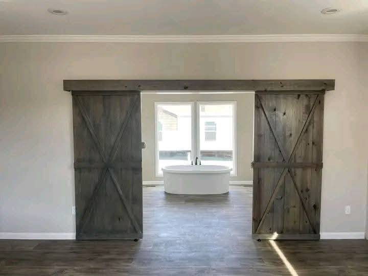 Wooden barn doors open to reveal a modern, freestanding bathtub beneath a large window, allowing natural light to fill the minimalist bathroom.