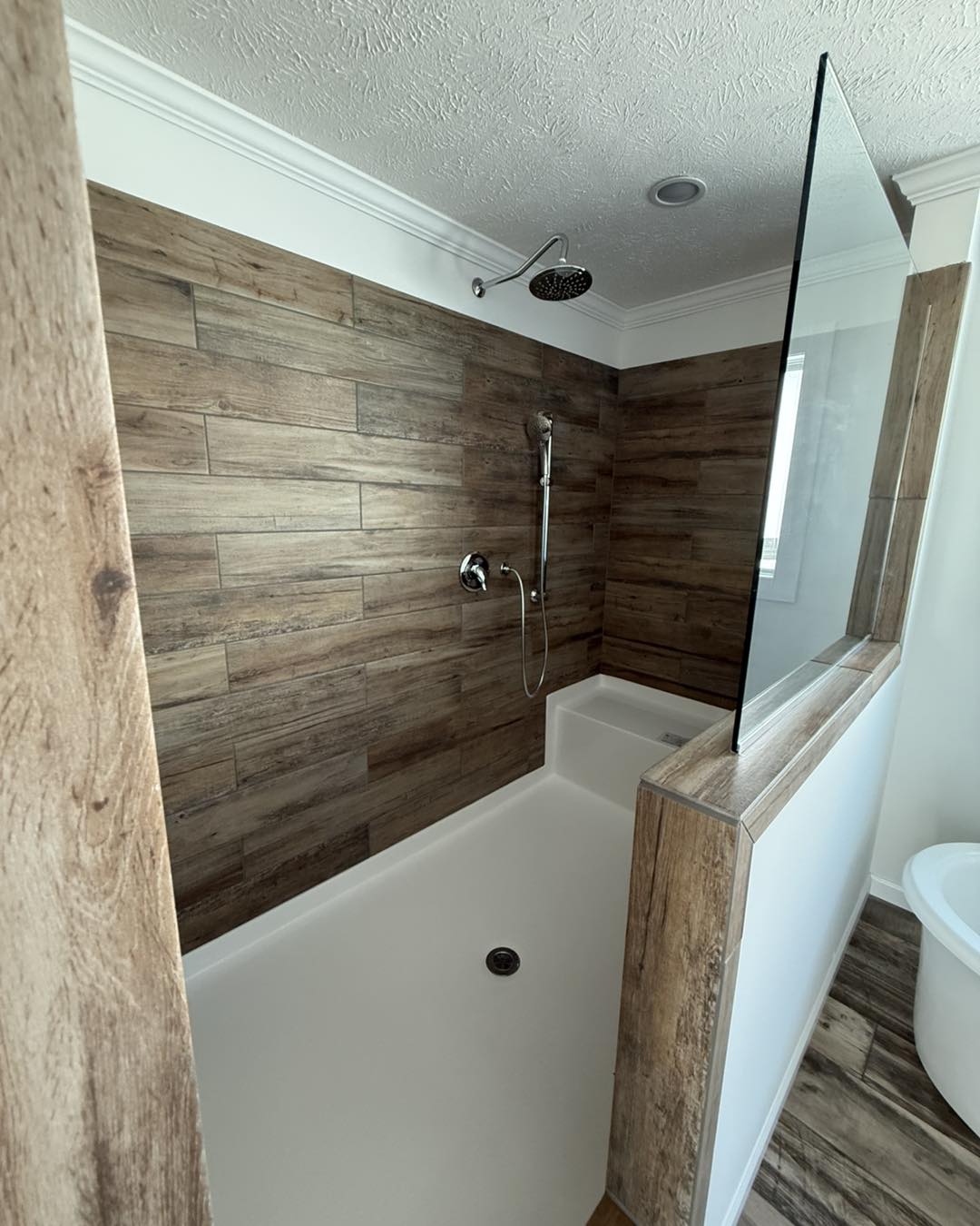 Modern bathroom with a wood-textured tiled shower, glass panel, and overhead showerhead. The atmosphere is sleek and minimalistic.