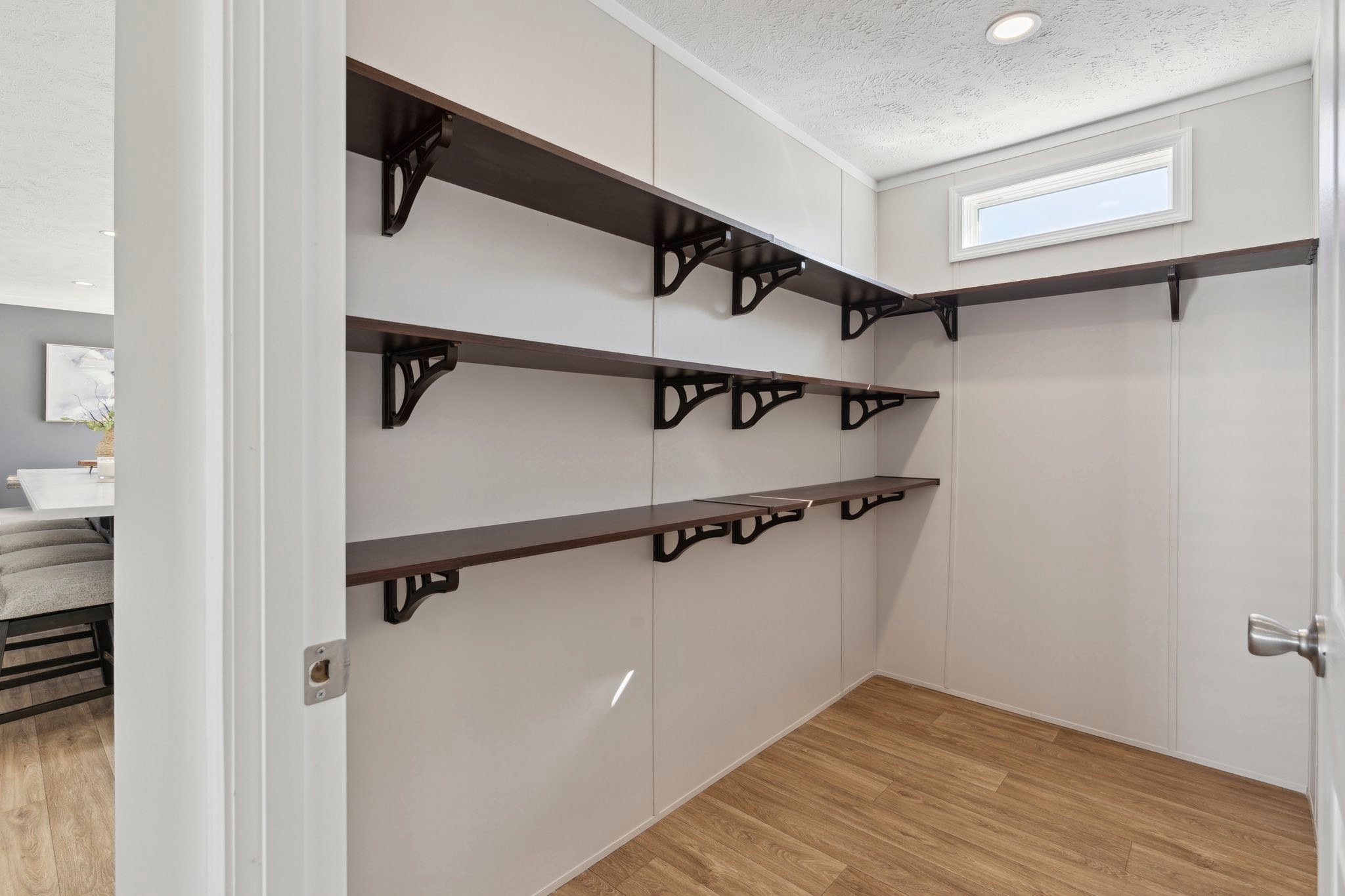 Spacious pantry with five dark wooden shelves on white walls, wooden floor, and a small window. Adjacent dining area visible with chairs and table.