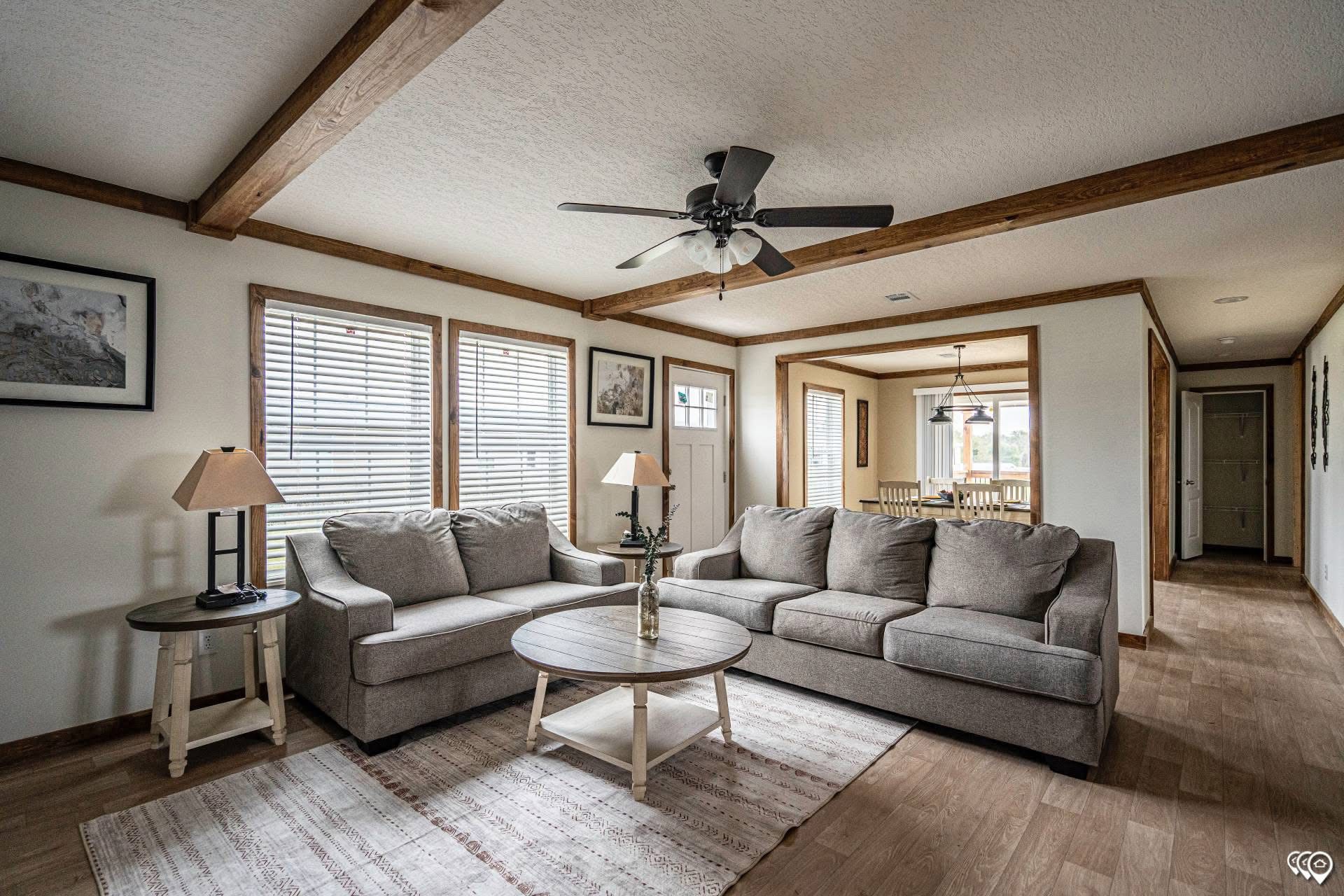 Spacious living room with a large gray sectional sofa, round coffee table, ceiling fan, and wood beam accents. Bright windows and cozy ambiance.