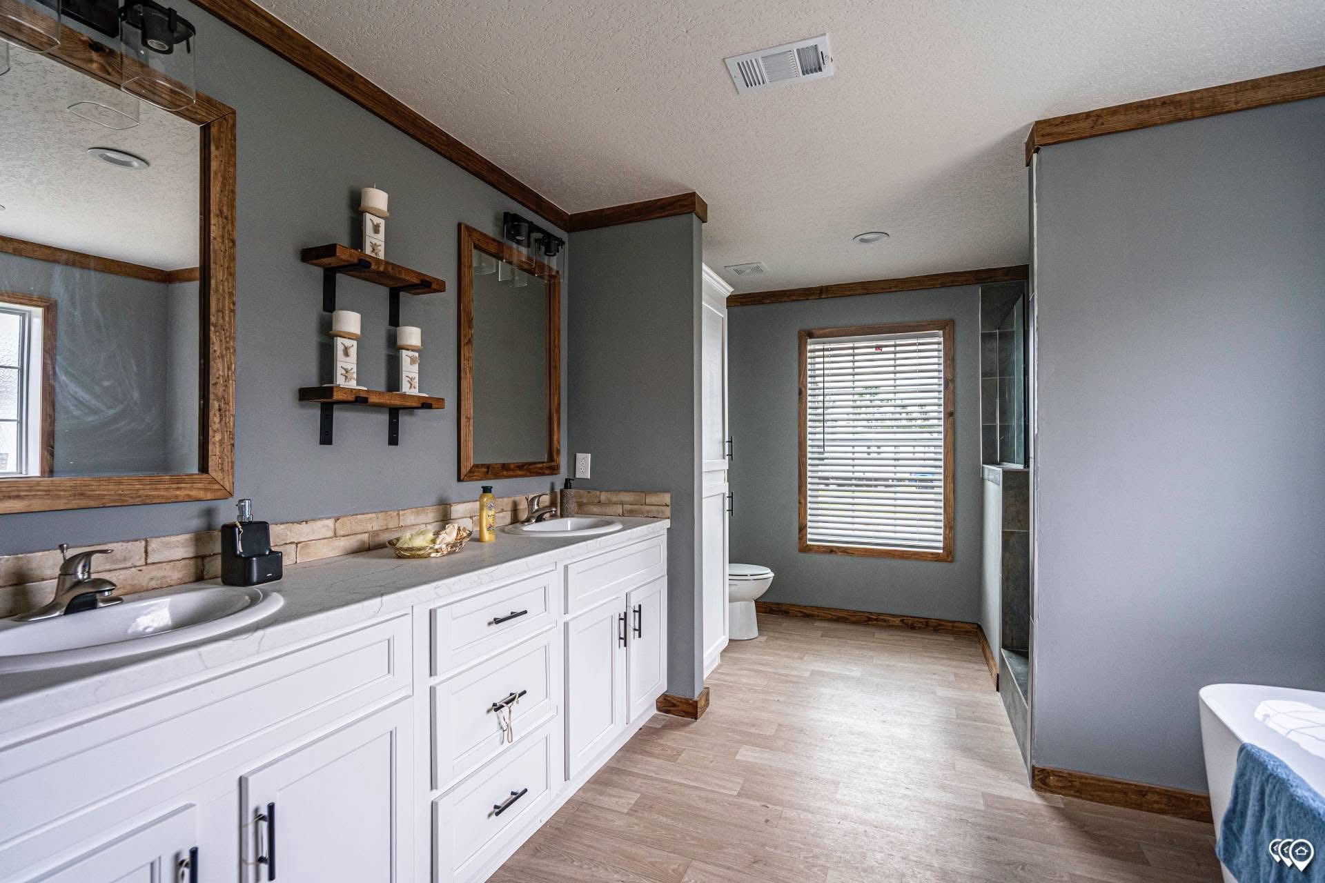 Stylish bathroom with gray walls, wood accents, and dual sinks. Large mirrors hang above the white vanity, with shelves holding candles. Bright, spacious, tranquil.