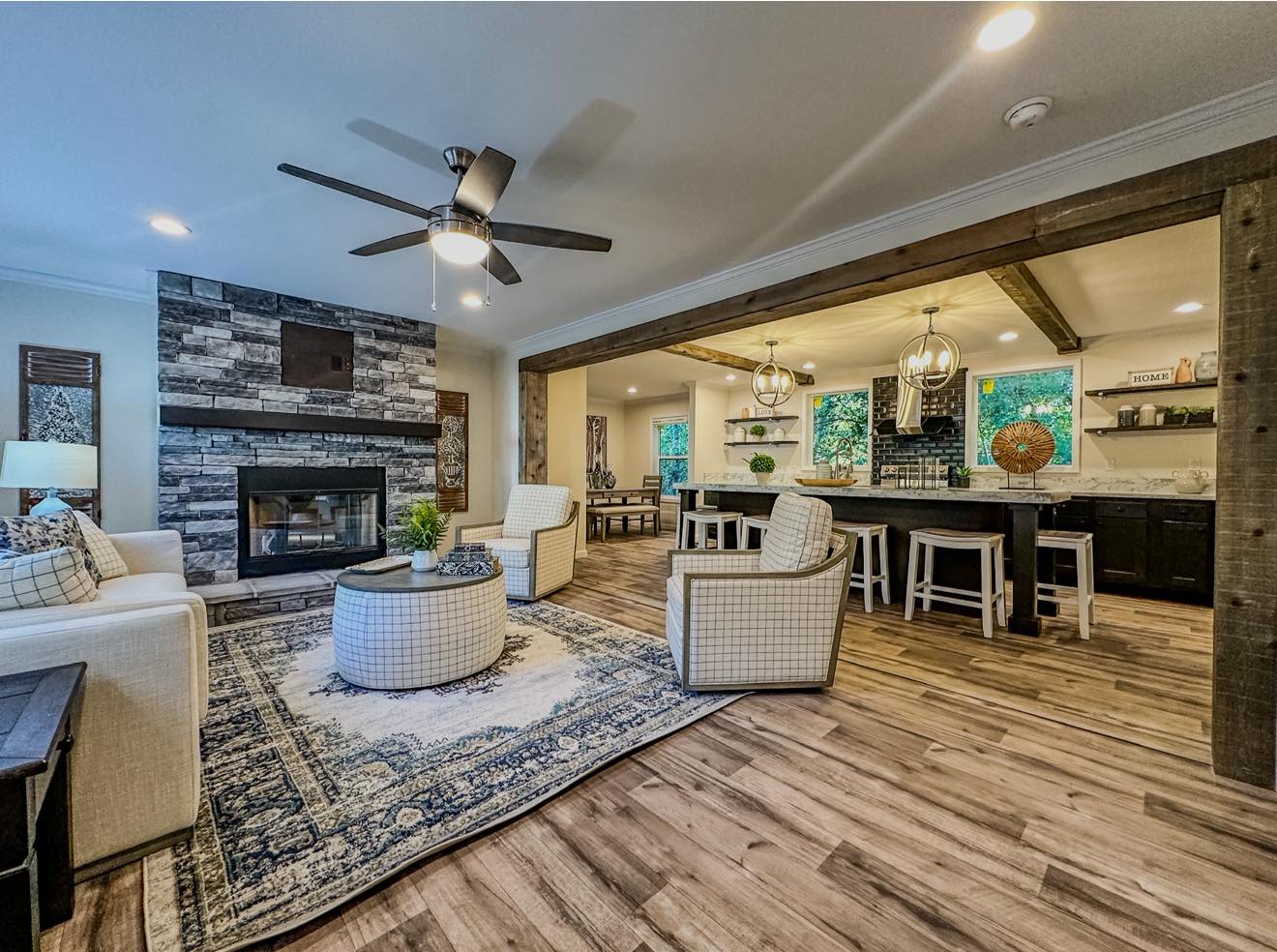 Stylish modern living room with stone fireplace, white sofas, and checkered chairs. Open layout leads to kitchen with bar stools and pendant lights.