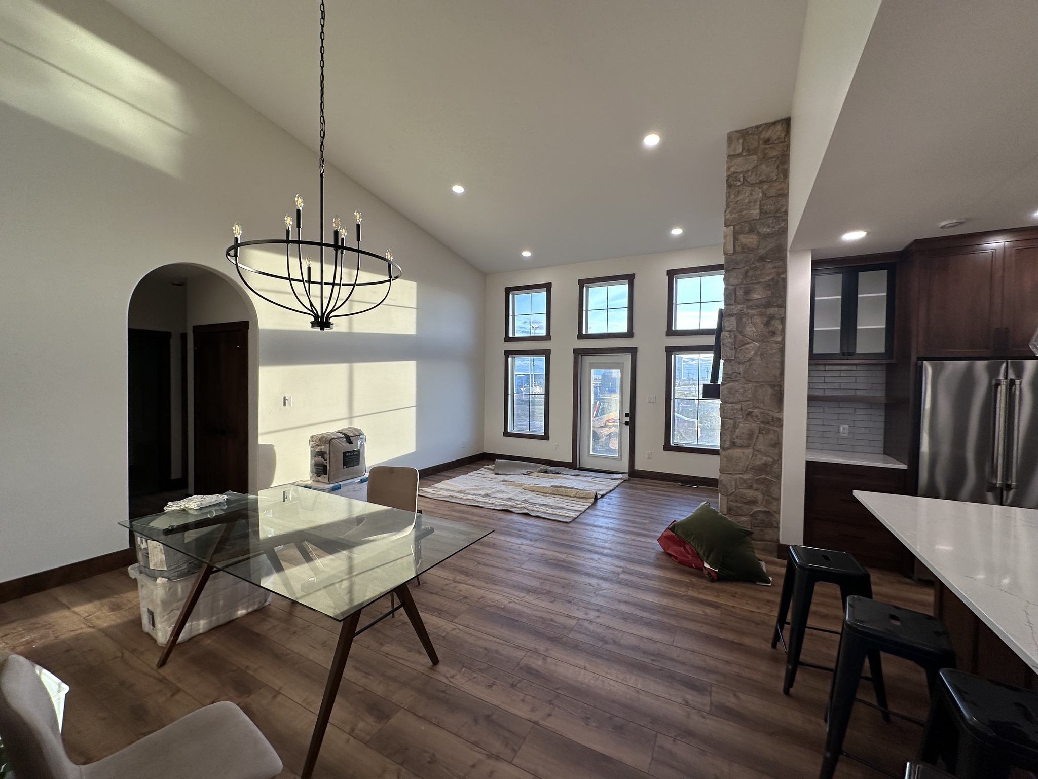 A modern living space with wooden floors, large windows, and a stone accent wall. A glass dining table, chandelier, and kitchen area enhance the cozy atmosphere.