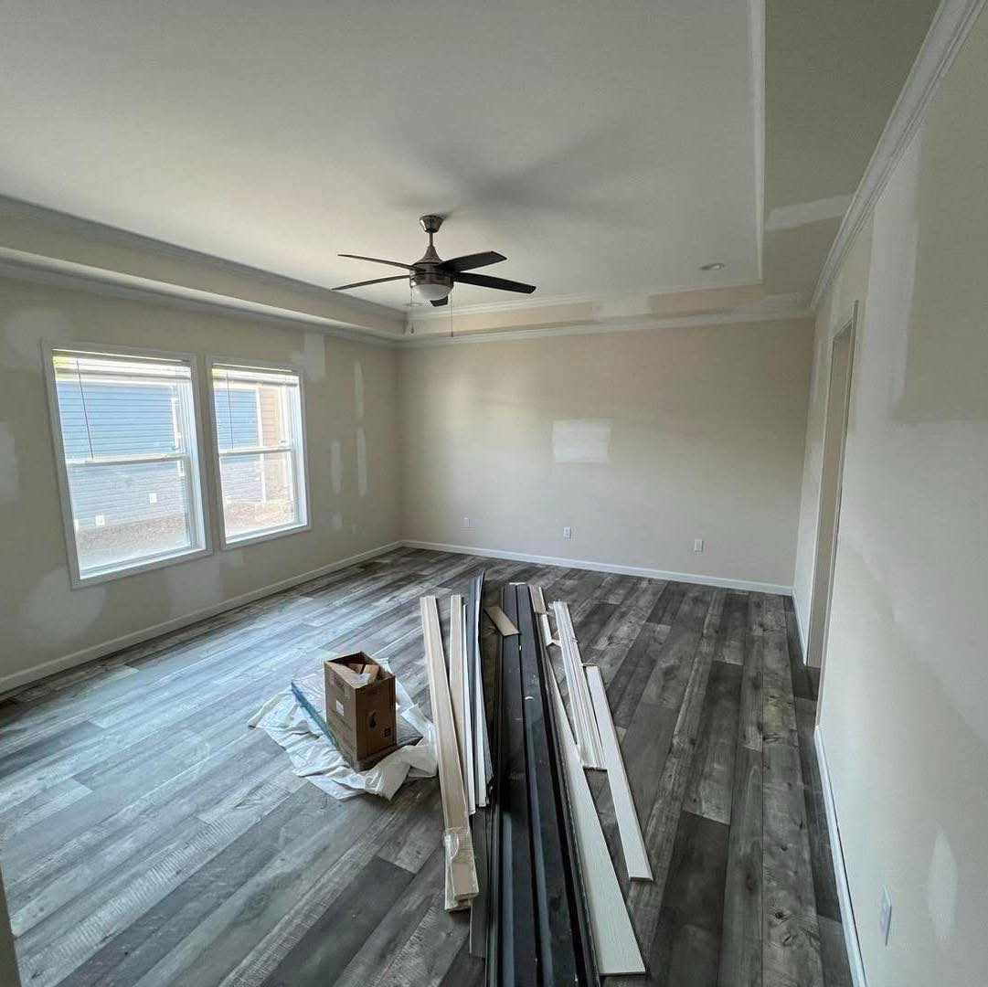 Spacious room with gray laminate flooring, beige walls, a black ceiling fan, and large windows. Construction materials are on the floor, conveying a work-in-progress vibe.