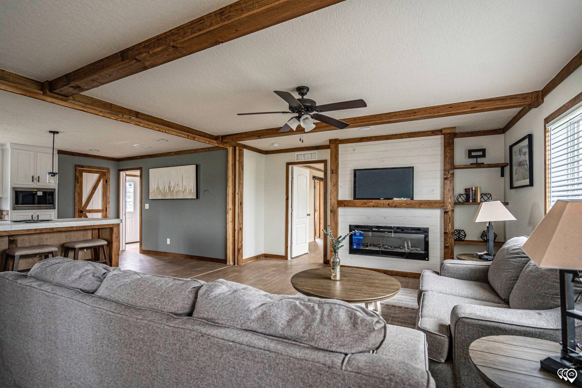 Spacious living room with exposed wooden beams and neutral tones. Features a gray sofa, round coffee table, fireplace, and cozy armchairs. Bright, inviting atmosphere.