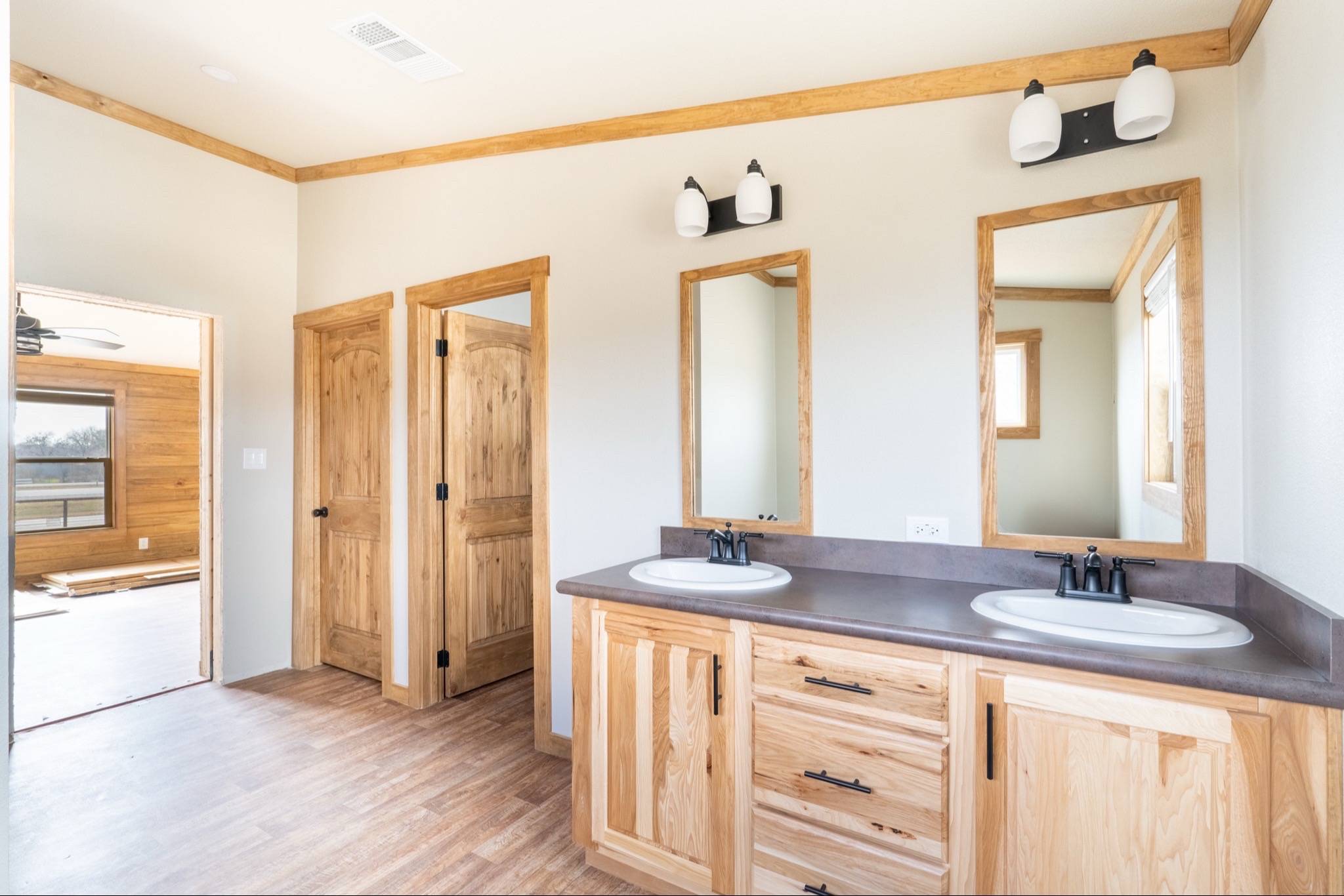 Spacious bathroom with wood accents featuring two sinks with dark faucets, matching mirrors, light fixtures, and wooden cabinetry. Warm, rustic tone.