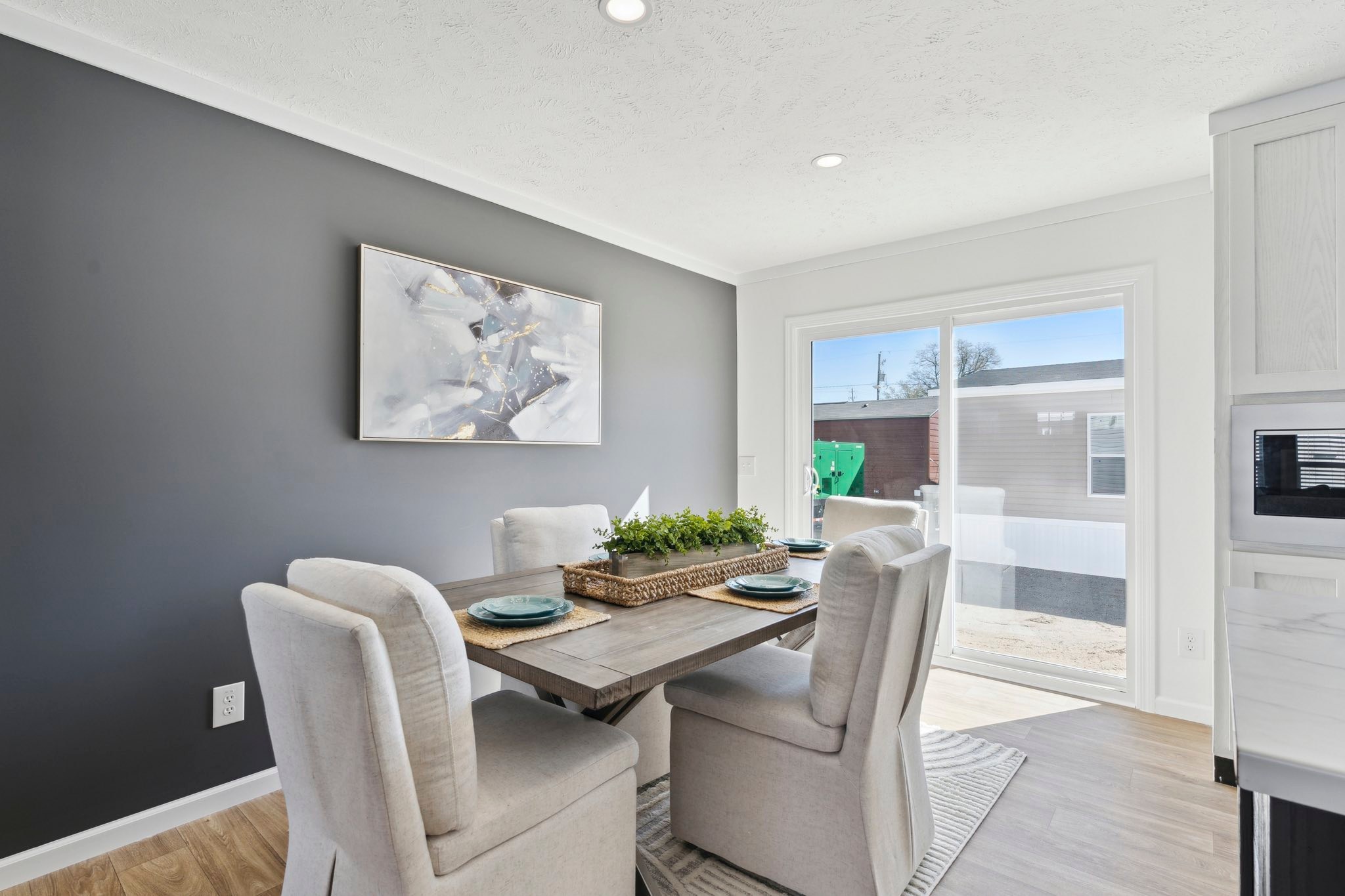 Contemporary dining room with a gray accent wall, abstract painting, and four cushioned chairs around a wooden table. Sunlight streams through glass doors.