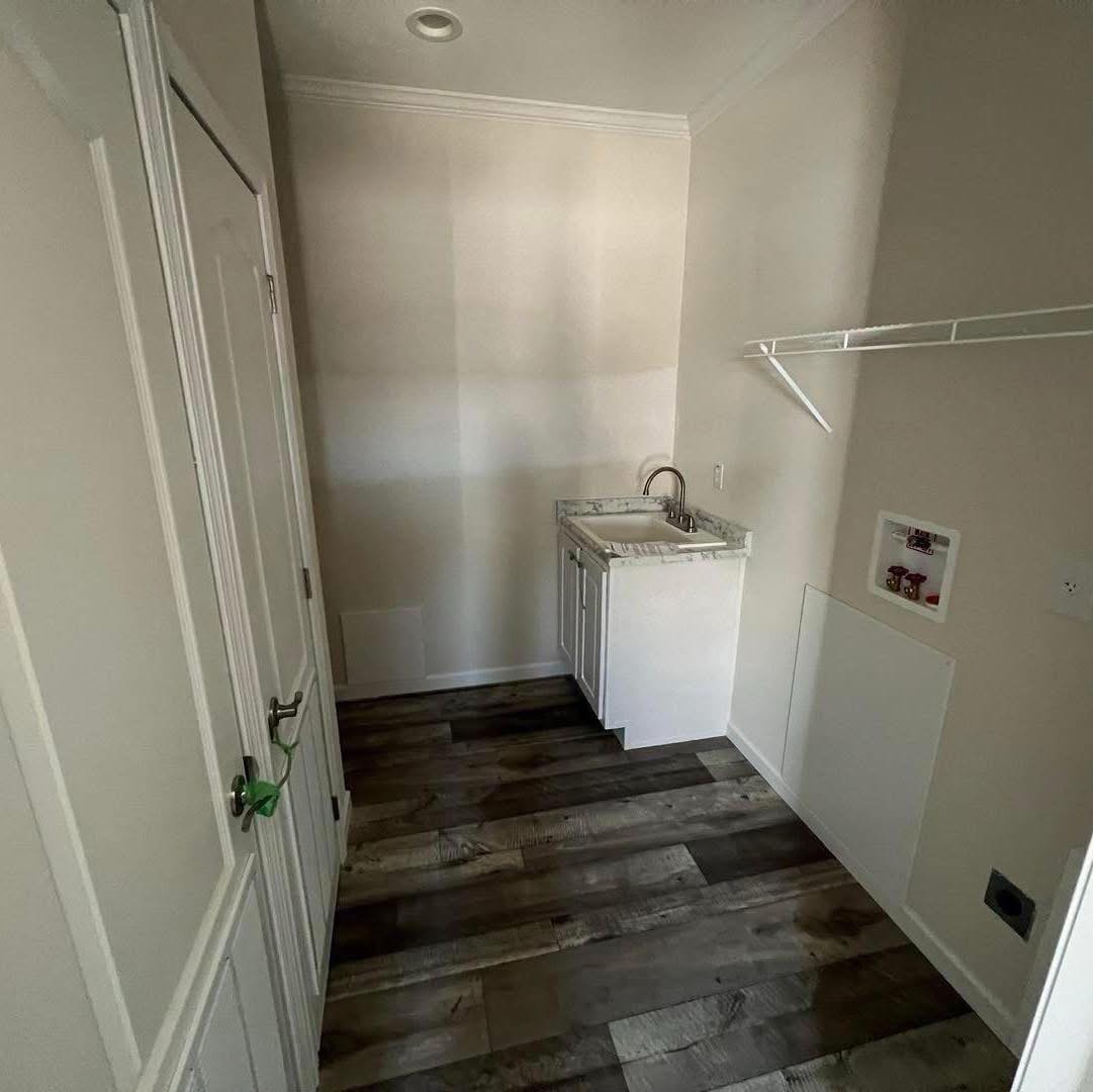 A small laundry room with a light marble countertop and sink, surrounded by white walls. Dark wood flooring adds contrast. A wire shelf is above, and a utility panel is on the wall, conveying a clean, functional space.