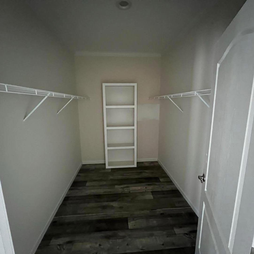 Empty walk-in closet with two white wire shelves on both sides and dark wood flooring. A tall white shelf unit is at the back wall. Bright, minimalistic space.