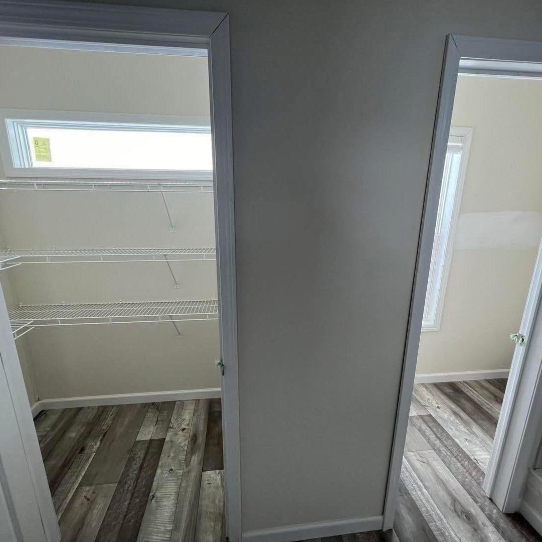 Two adjacent closets with white doors open to reveal a pantry with wire shelving on the left, and an empty space with a window on the right. Sunlight filters in, highlighting the wood-patterned flooring.