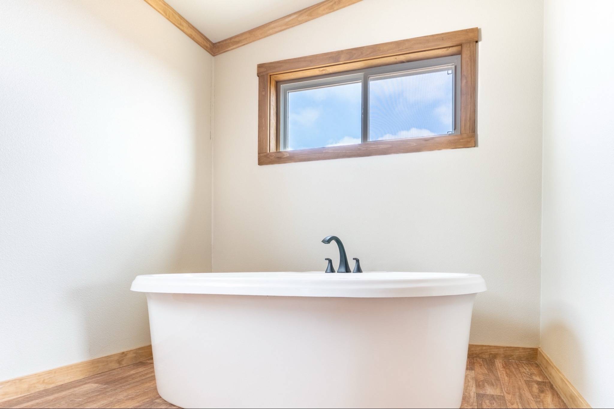 Minimalist bathroom with a white freestanding bathtub on wooden flooring. A window with wood trim above fills the space with natural light, creating a calm atmosphere.