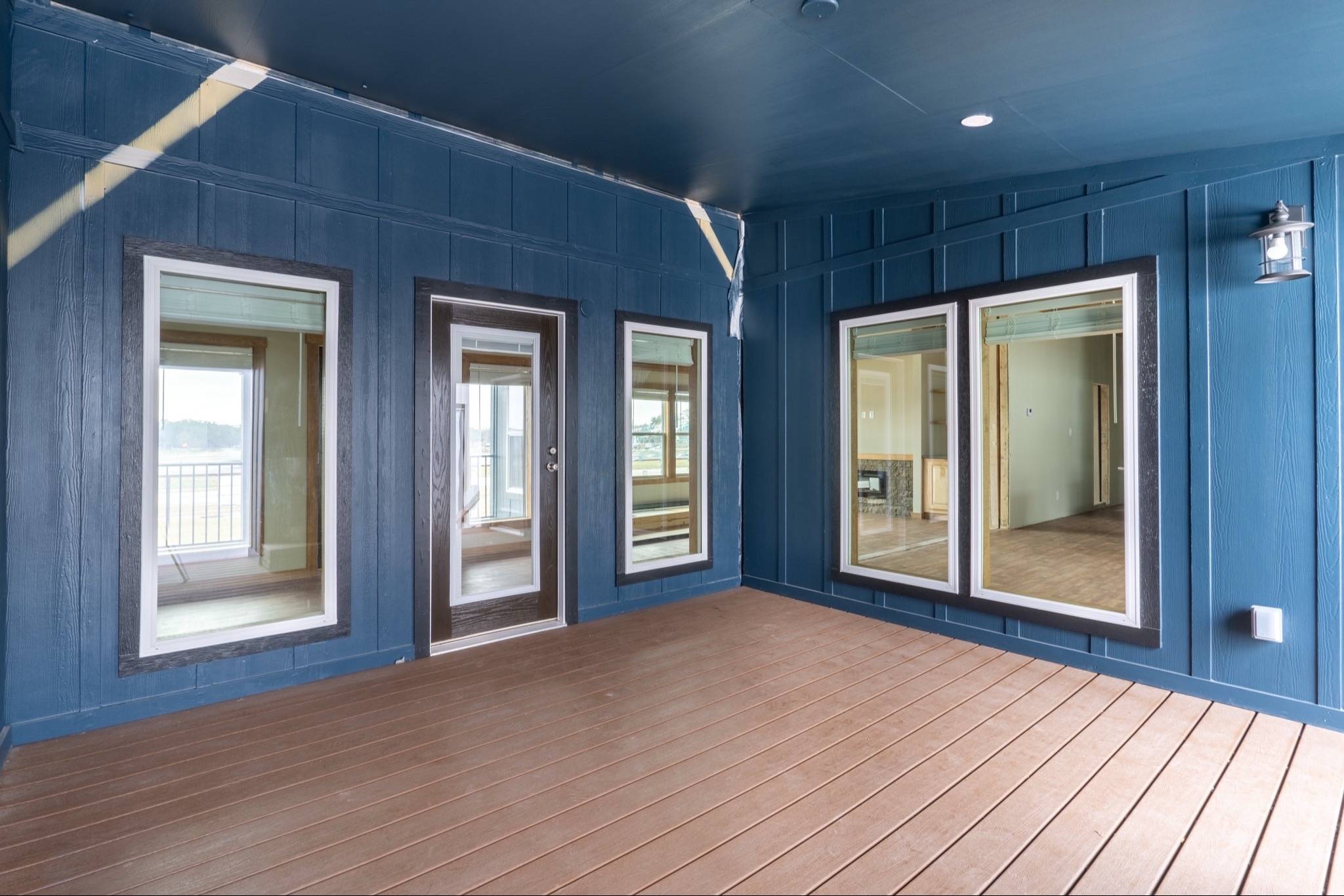 Covered porch with blue wooden walls, large windows, and a glass door. The floor is brown wood. Bright natural light gives a welcoming feel.