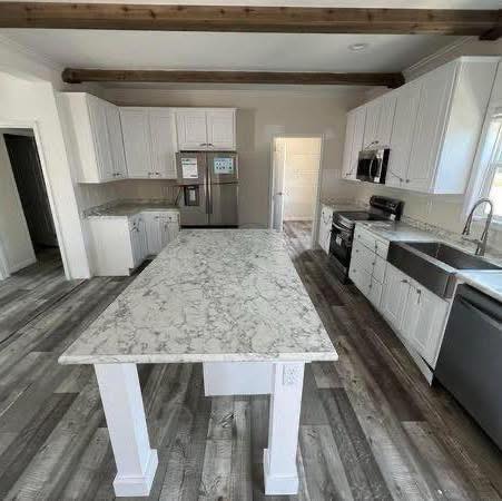 Spacious kitchen with a large marble island, white cabinets, stainless steel appliances, and wooden beams. Grey wood flooring adds a modern touch.
