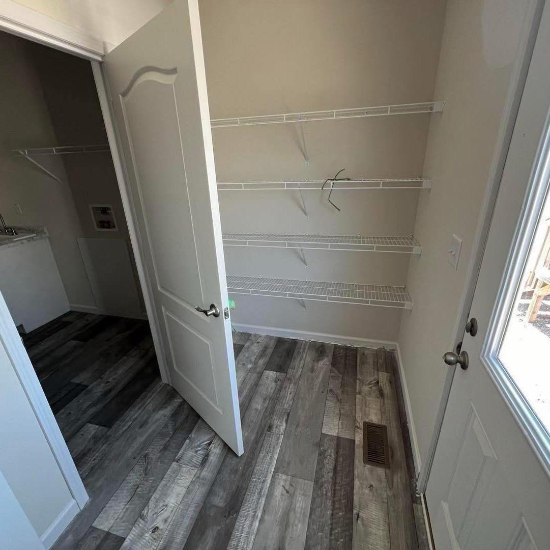 A small, empty pantry with light gray walls, wooden flooring in varied shades, and white wire shelving on two sides. A slightly open door leads to another room.