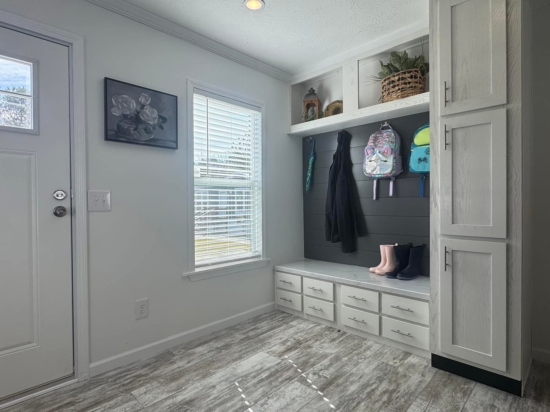 A bright mudroom with white cabinets and drawers, backpacks, coats, and boots. A window lets in light, highlighting the floral artwork and plants. Cozy and organized.