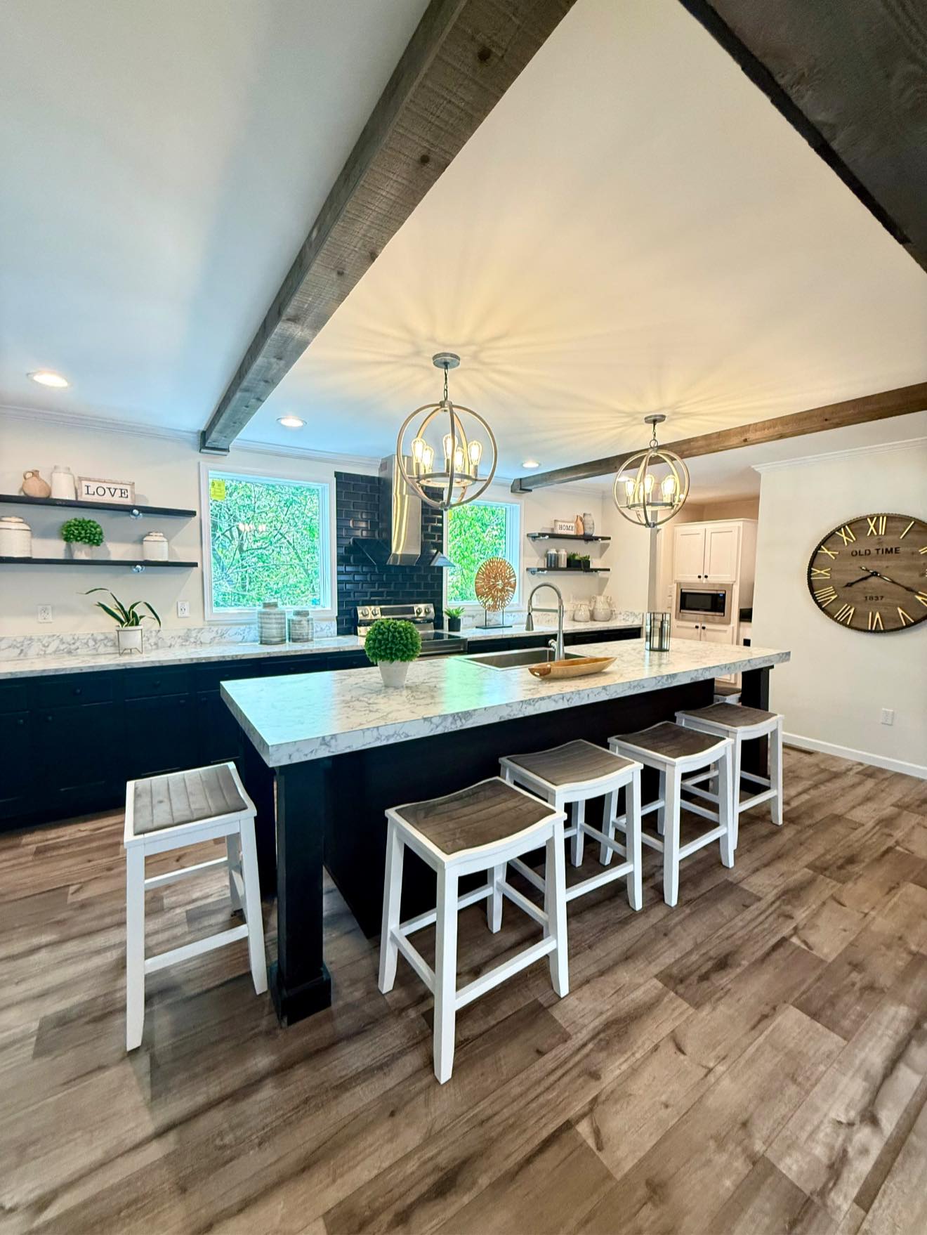 Modern kitchen with a large marble island, wooden stools, and orb pendant lights. Shelving with décor and large wall clock create a cozy, stylish atmosphere.