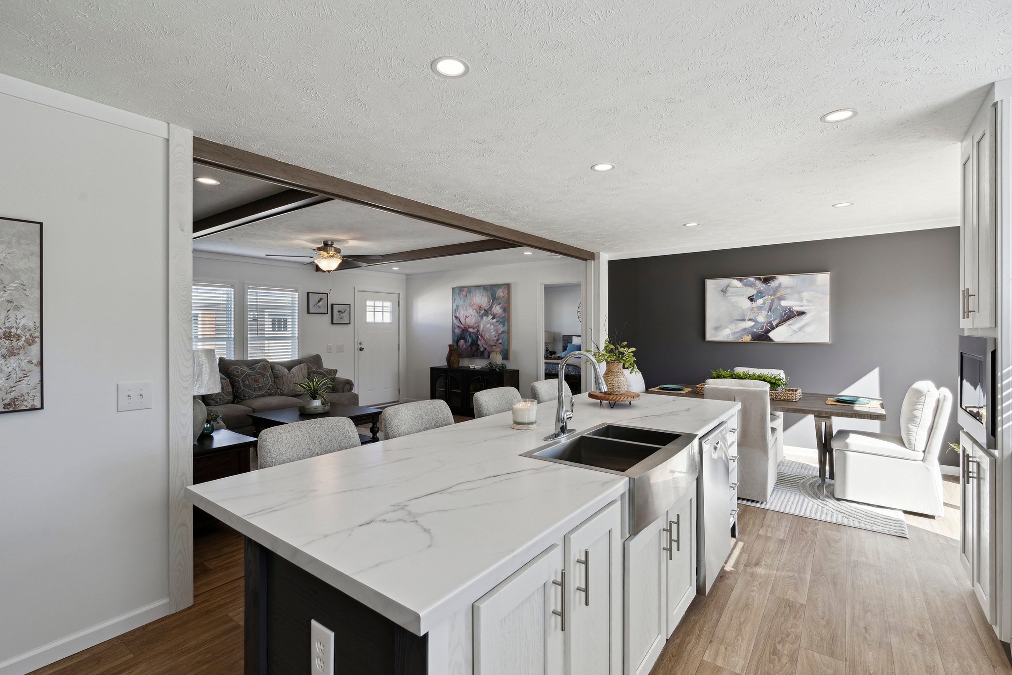 Modern open-concept kitchen and living area with white cabinets, marbled countertop, wooden floor, elegant dining table, and cozy gray sectional sofa.