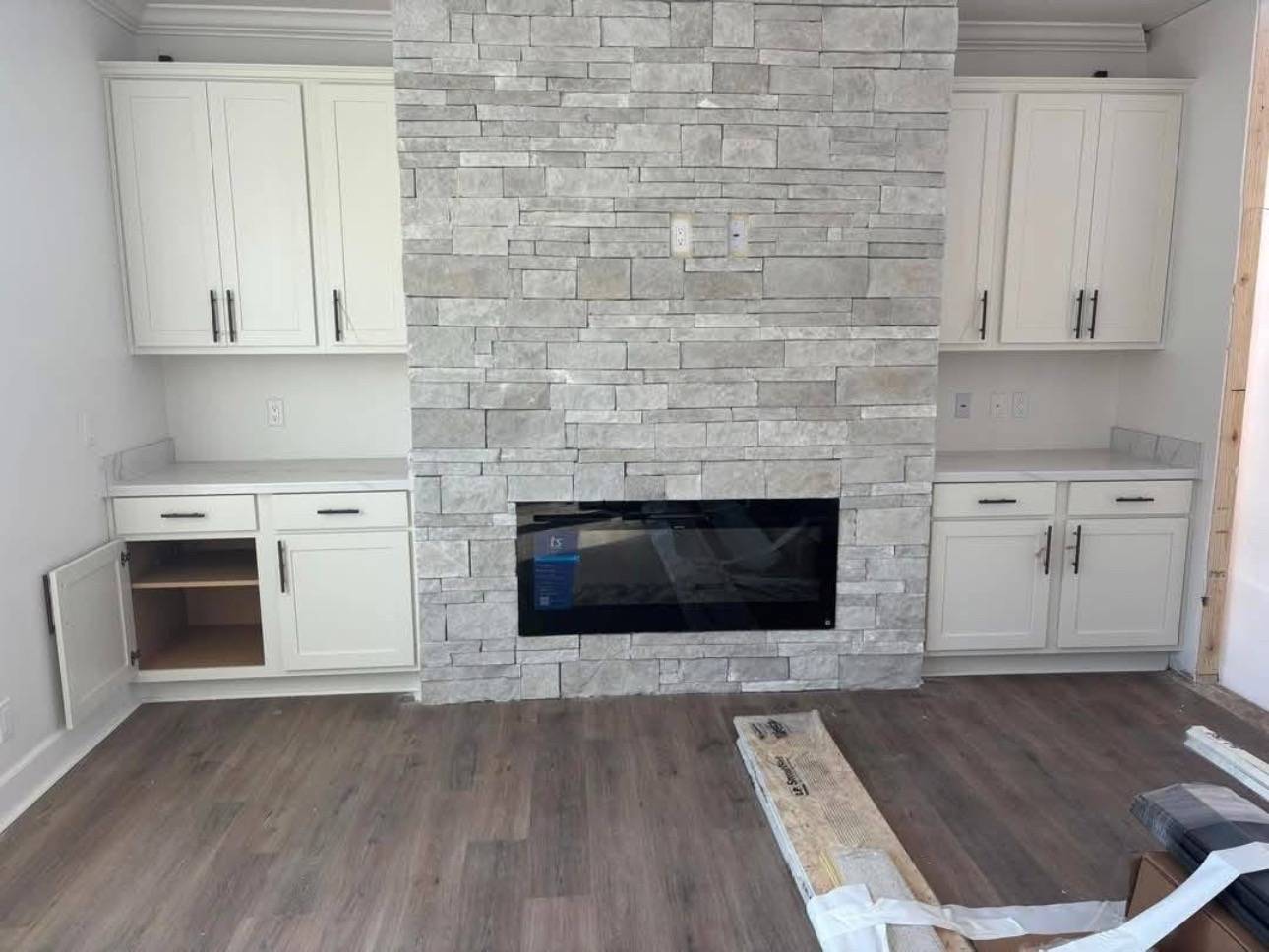 A modern living room with a stone fireplace centered between two white cabinets. Light wood flooring adds warmth. The open cabinet suggests ongoing work.