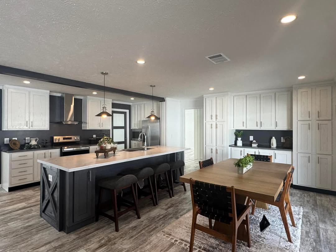 Modern kitchen with white cabinets, a dark island, and pendant lights. A wooden dining table with black chairs sits on a patterned rug, creating a cozy atmosphere.