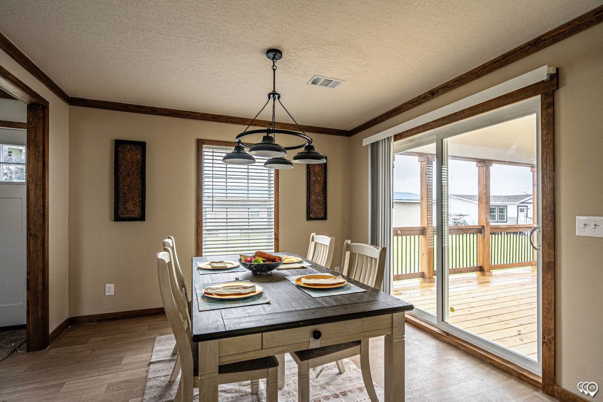 A cozy dining room with a rustic wooden table set for four, framed by large windows and a glass door leading to a deck. Warm tones and soft lighting create a welcoming atmosphere.