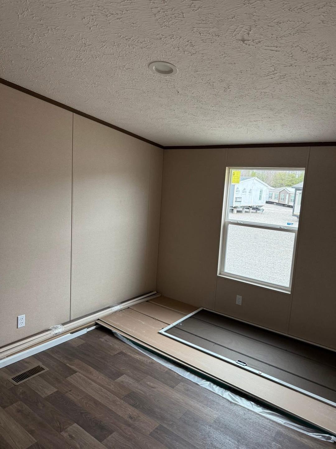 A small, unfurnished room with beige walls and dark wood flooring. A window on the right shows an outdoor view of houses. Construction materials lie on the floor.