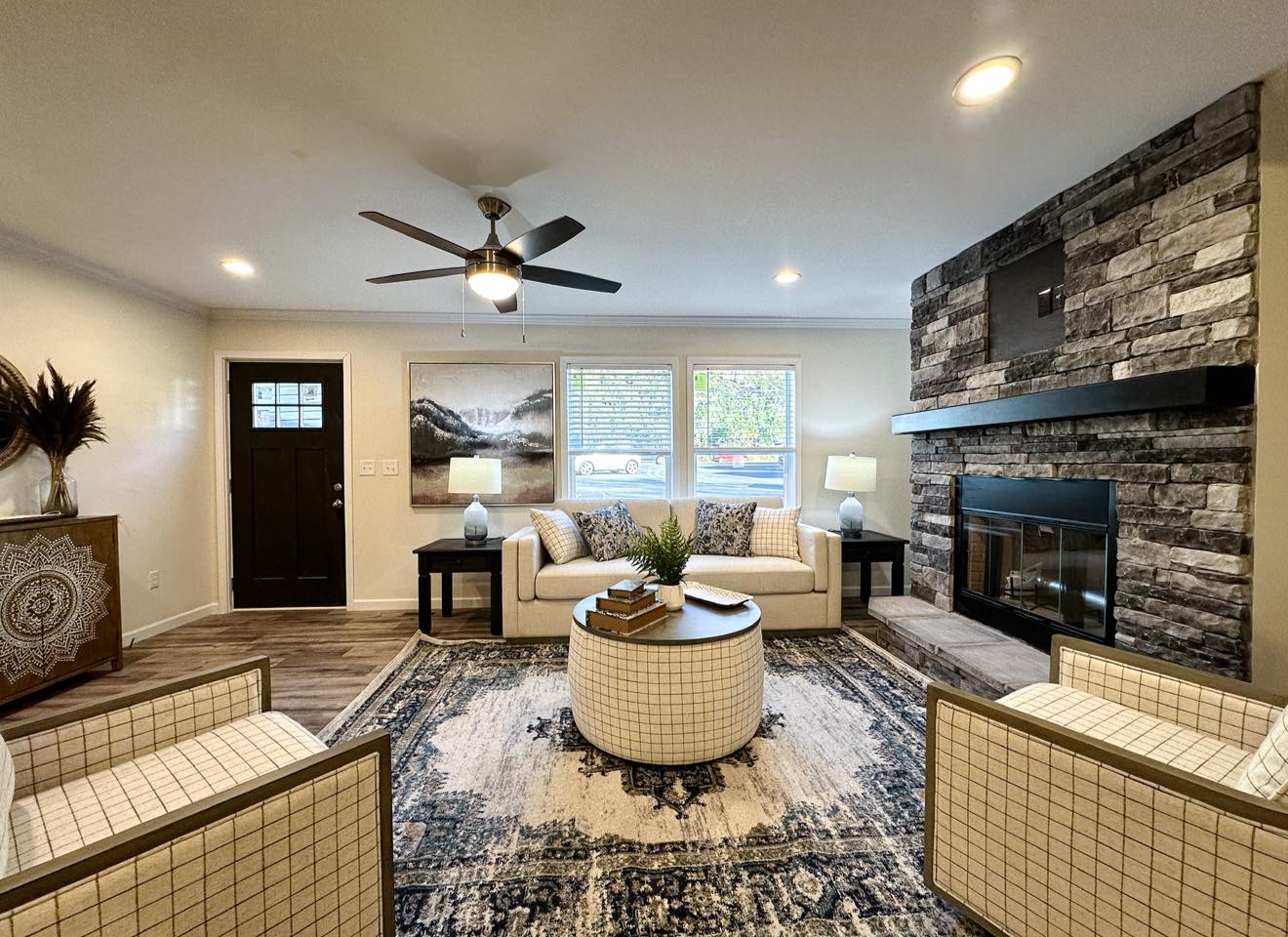 Modern living room with stone fireplace, ceiling fan, and patterned rug. Neutral tones with a round coffee table, two armchairs, and cozy sofa. Calm ambiance.